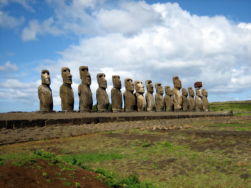 Fifteen huge monolithic stone statues of partial human figures with big heads lined up against blue sky.