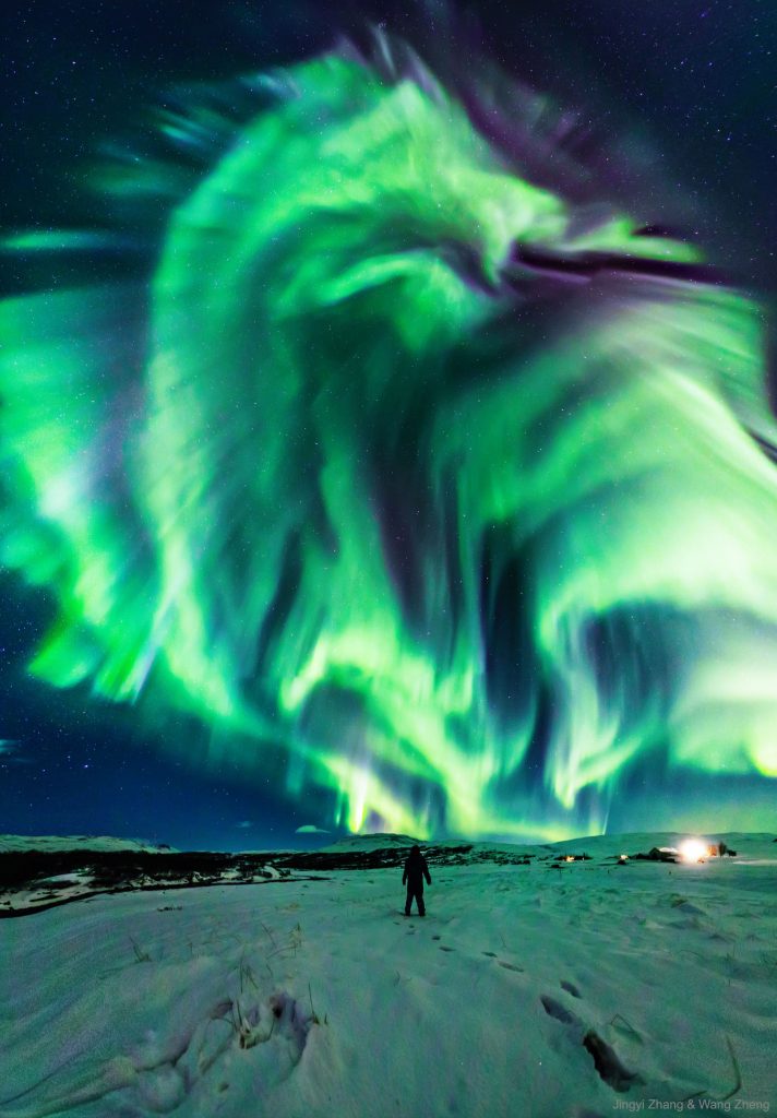 Vast green dragon-like aurora nearly filling the sky with a person standing on snowy lanscape.