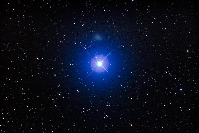 A single brillian bluish star on a dense star field. Above it is a faint oval smudge of light.