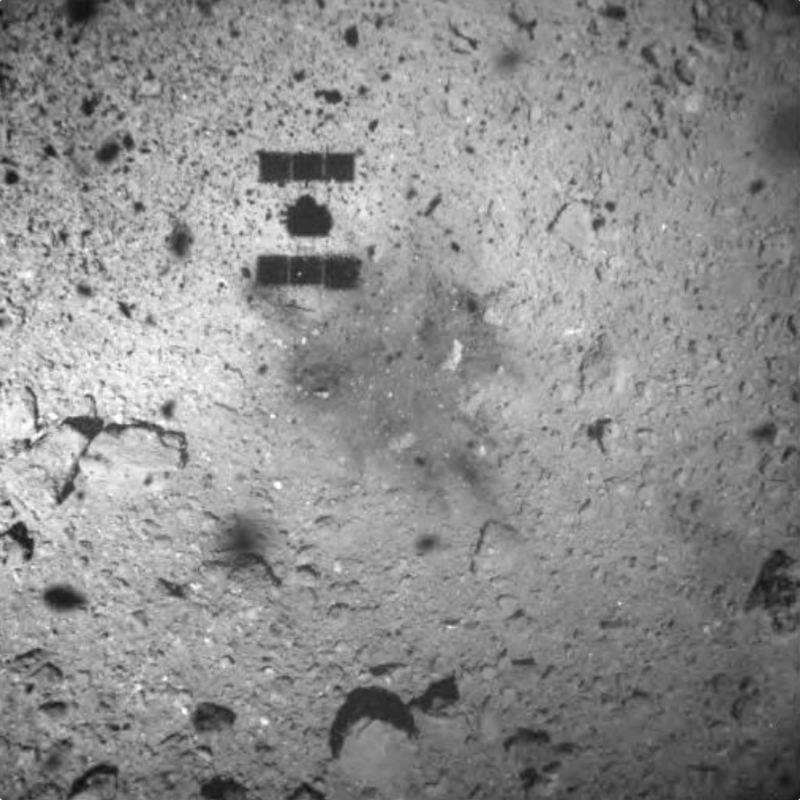 Spacecraft shadow, and a dark irregular spot, on the surface of a rocky gray asteroid.