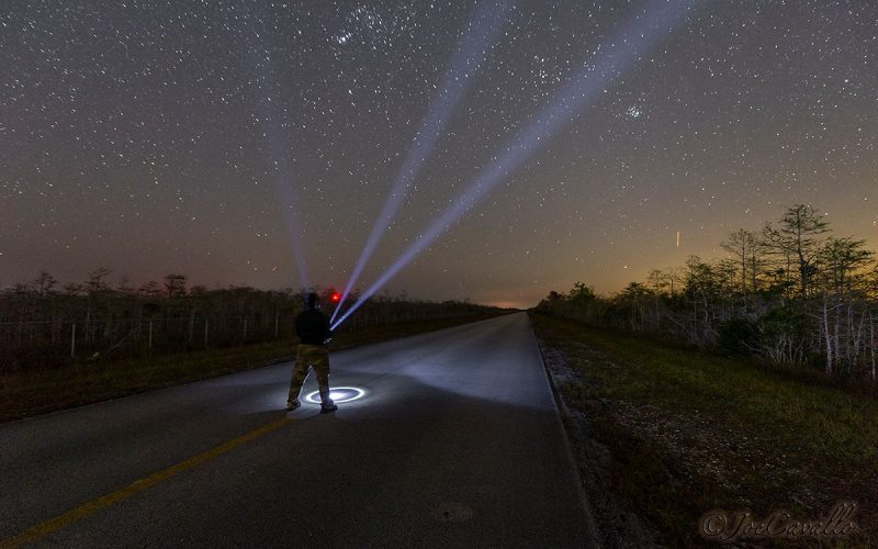 Man standing on a highway shining a flashlight into a starry sky.