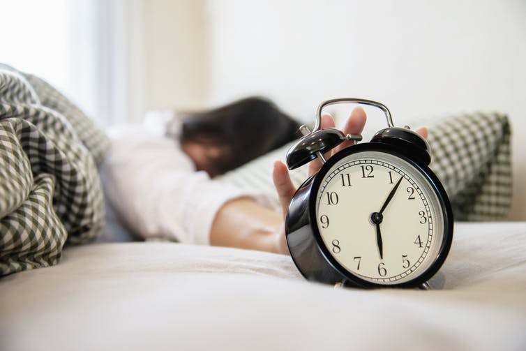 Life: Sleepy woman reaching for alarm clock with bells on top set to 6:07.