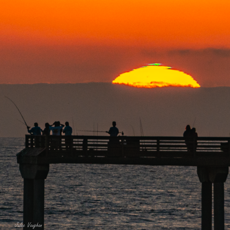 Setting sun, mostly below the ocean horizon, with short green upper rim, and a fishing pier in the foreground.