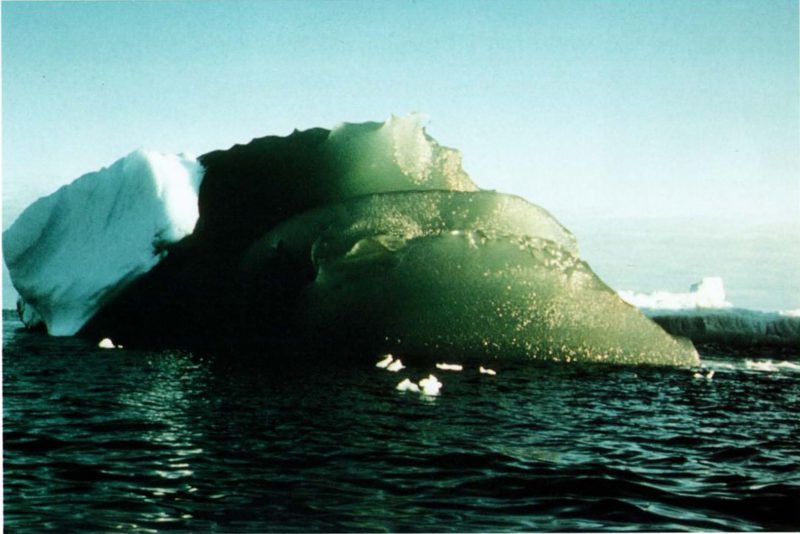 Green iceberg with blue iceberg behind it both floating in dark sea.