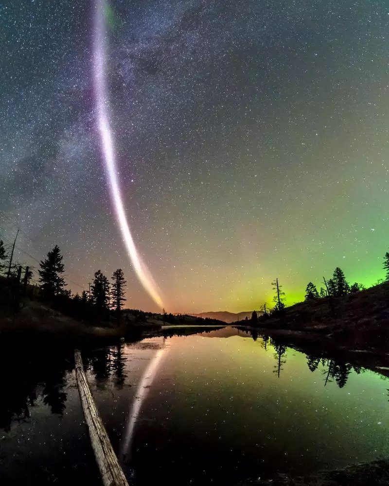 Curved pink beam shoots into sky over lake with green aurora in distance.