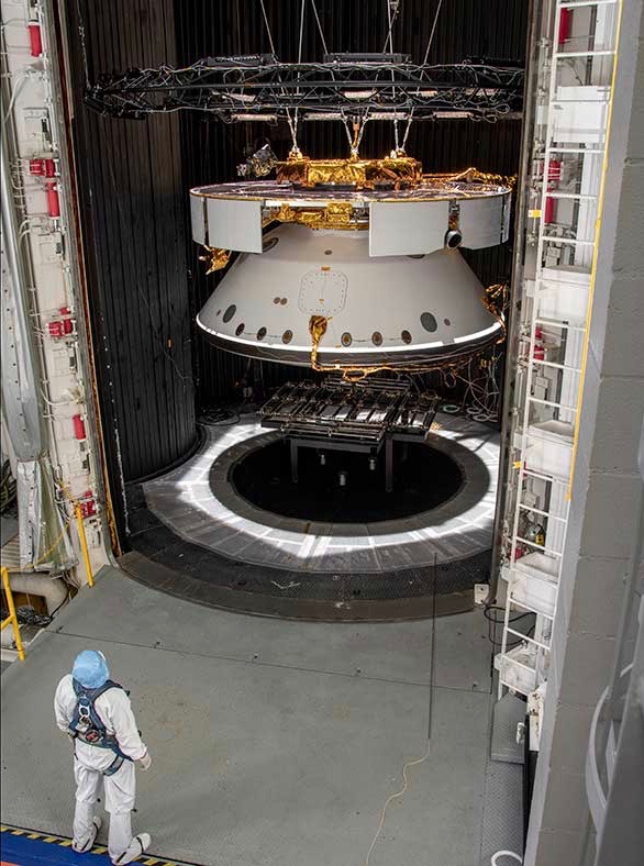 A man in a clean suit stands looking at a round spacecraft, conical below.