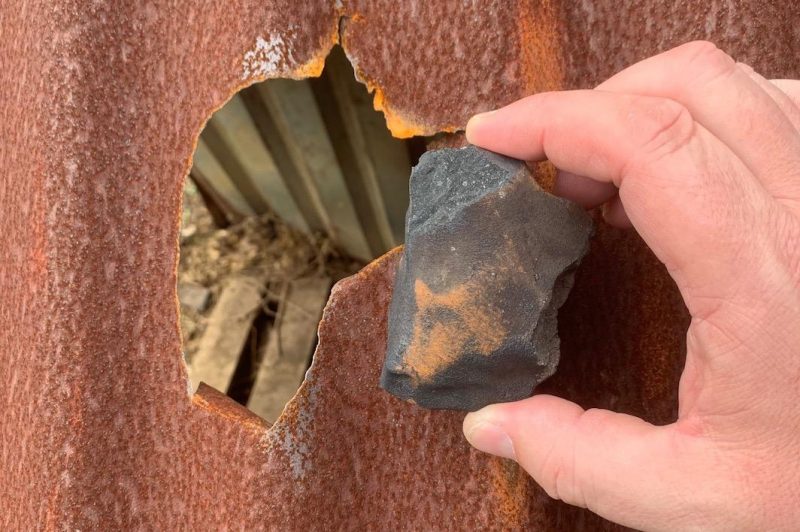 Hand holding a small, dark, irregular meteorite over hole in doghouse roof.