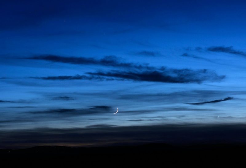 Twilight sky, clouds running left to right, very thin, low crescent moon, two bright dots.