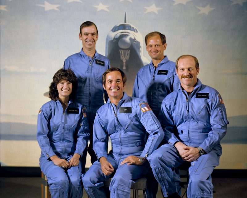 One woman and four men in blue flight suits, with space shuttle in background.