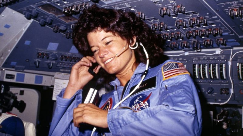 Woman with plentiful curly hair, wearing blue astronaut outfit and headphones, in spacecraft cockpit.