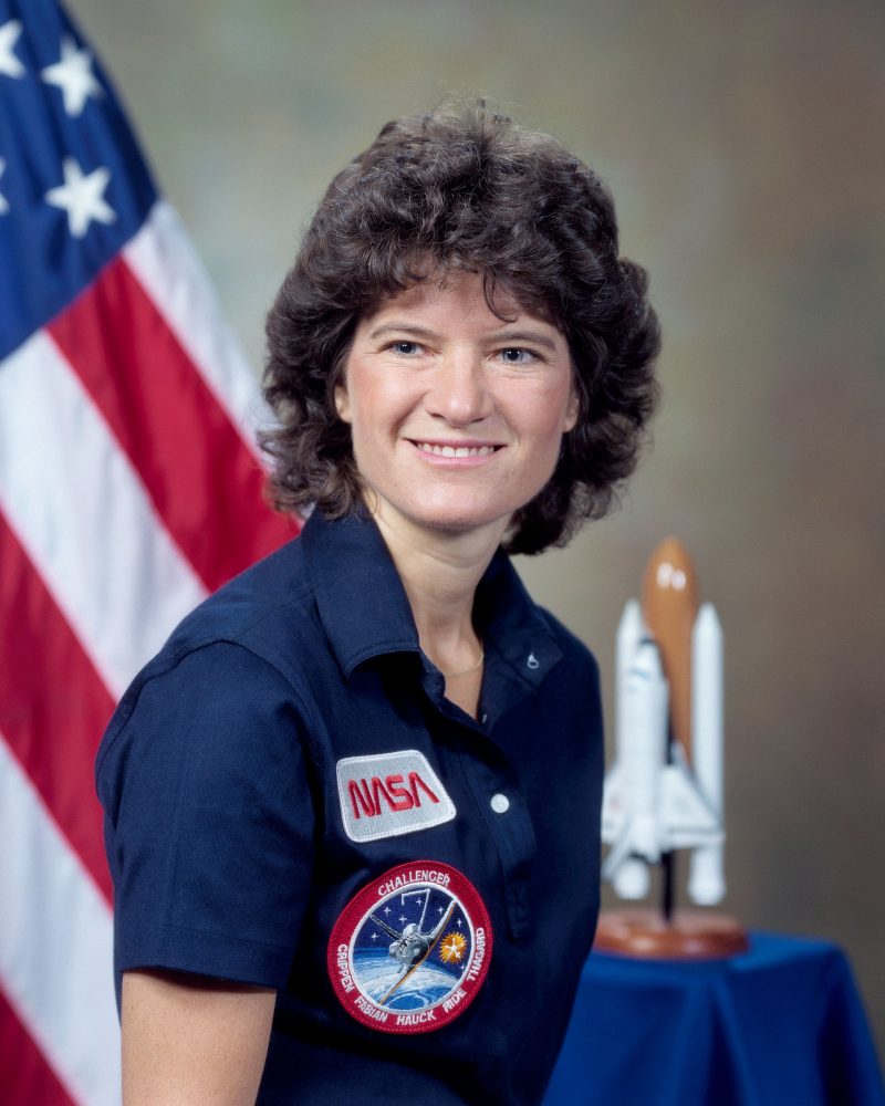 Smiling woman in dark shirt with NASA patches, with model of space shuttle and American flag.