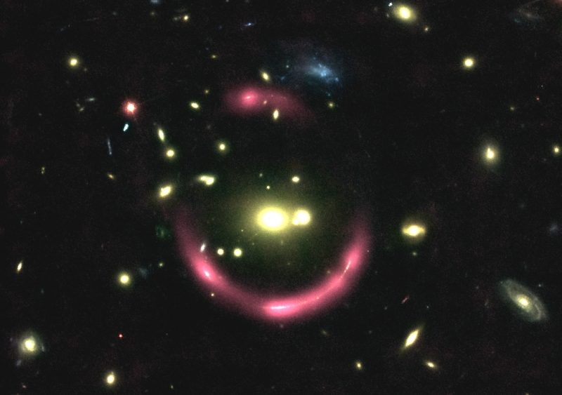 Fuzzy ring of pink with white nodes in it around 2 yellow galaxies, with other galaxies in background.