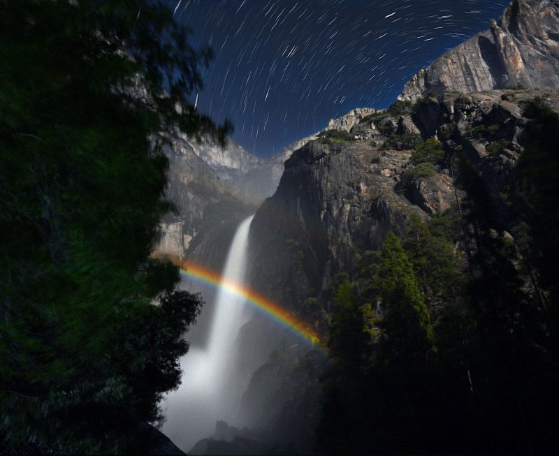 Rainbow over a rocky waterfall with star trails in night sky above.