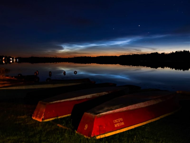 Colorful nighttime image of upturned boats on a waterfront, with noctilucent clouds shining overhead.
