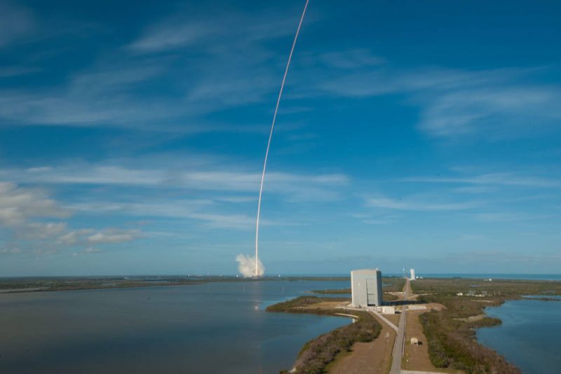 Distant launch. Small puff of steam at base of curved vertical white line in a blue sky.