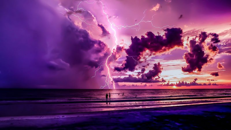 Two people standing on the beach with sunset/ twilight colors and lightning striking the water far in the distance.