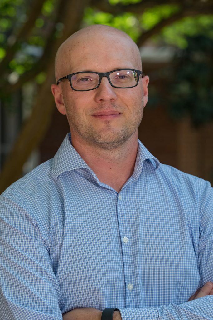 Satisfied looking bald young white man with glasses, blue checked shirt and folded arms.