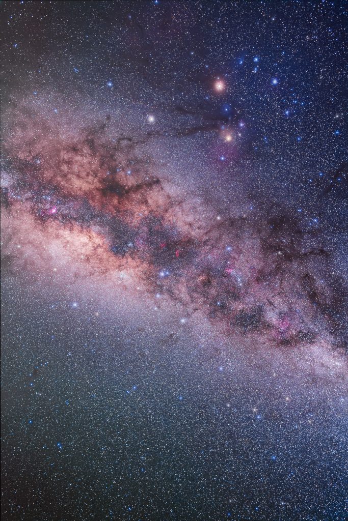 A rich star field showing the center of the Milky Way in nebulous red, white and pink, with dark dust lanes. Scorpius, Mars and Saturn appear with Antares and Mars bright red while Saturn is yellowish-white.