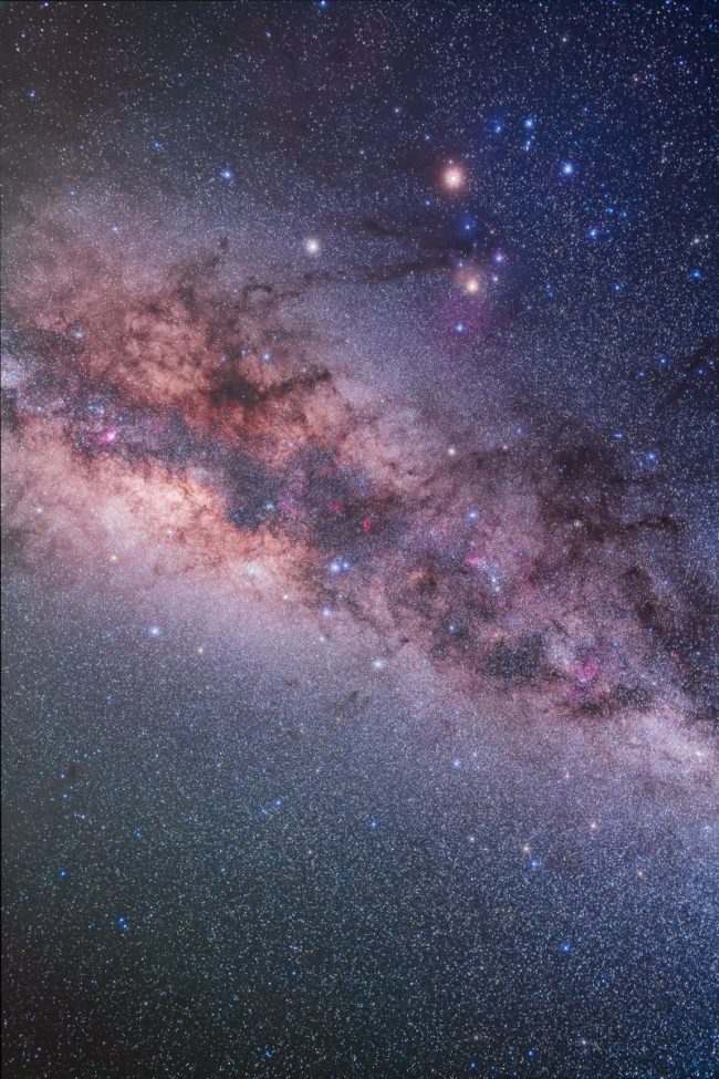 Very dense star field with Milky Way in nebulous colors nd dark dust lanes, with two large reddish dots.