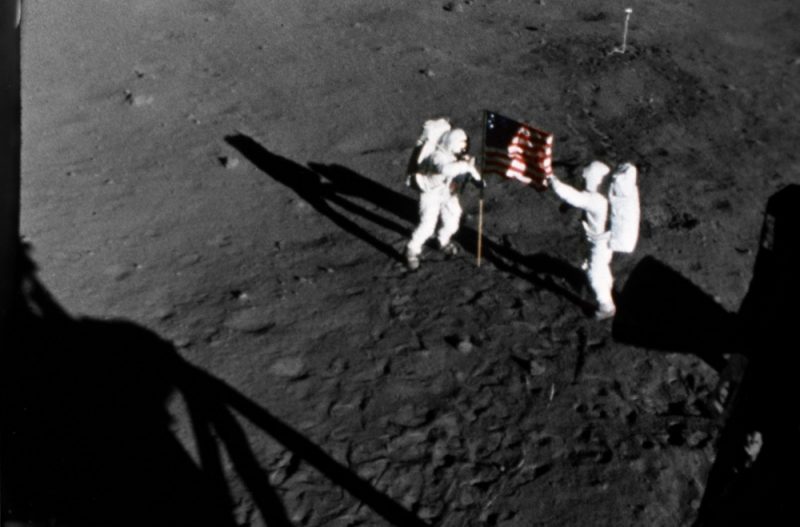 View from above of 2 astronauts in spacesuits deploying a US flag on the moon.