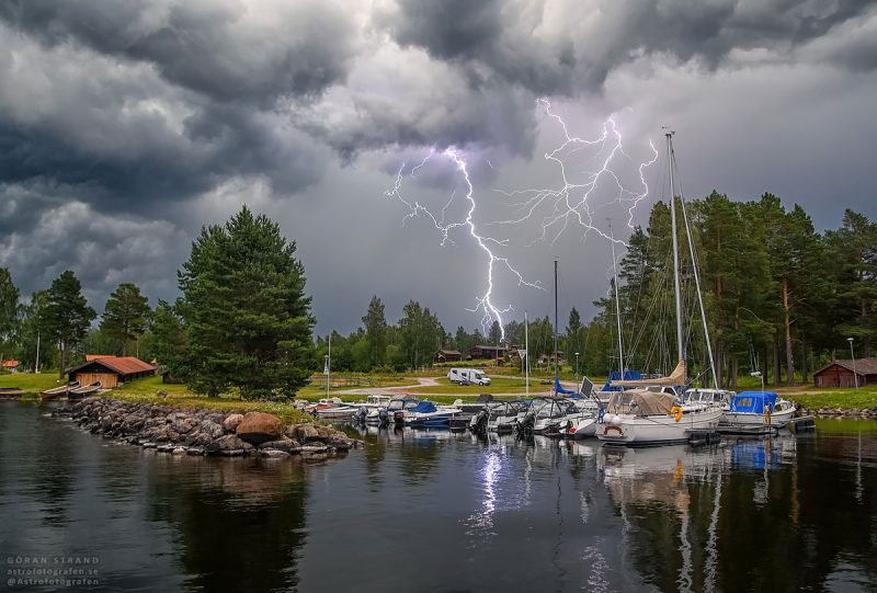 Jagged vertical streak of light from gray sky to ground behind inlet with docked sailboats.