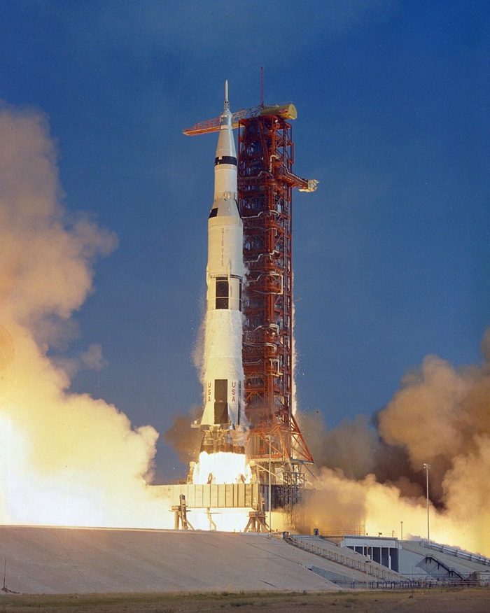 Rocket on a launchpad against a blue sky, with smoke billowing at its base and red gantry next to it.