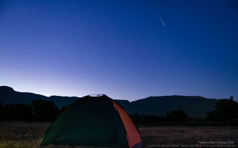 Bright meteor in a bluish sky, above a small dome-style tent and hilly horizon.