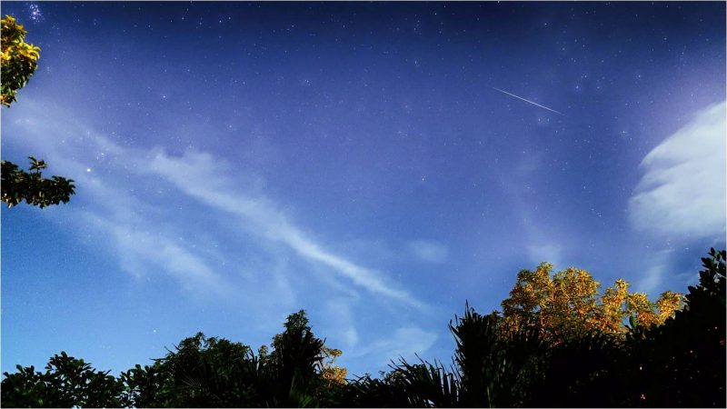 Bright meteor streaking along in a daytime-like bluish sky with some elongated white clouds.