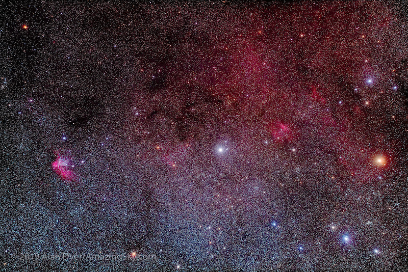 A bright star in the center, Delta Cephei, against a backdrop of fainter stars and wispy red interstellar clouds. To the left of the center star is a reddish nebulous feature, the Wizard Nebula. To the right of the star is another fainter red nebulous region, Sharpless 2-135. Right of Sharpless 2-135 is an orange star, Zeta Cephei.