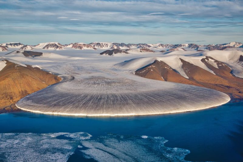 Wide flat round front glacier extending from between bare hills out into blue sea.