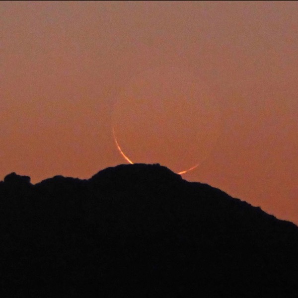 Thin yellow crescent, partly behind a wooded hill, in a dark orange twilight sky, with rest of moon showing dimly.