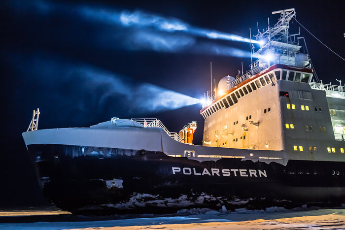 Bow end of very large ship with three searchlights shining forward.