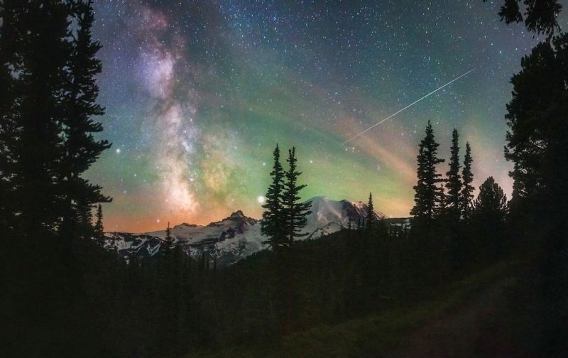 Dark mountains in distance, tall evergreens in foreground against sky with colorful phenomena.