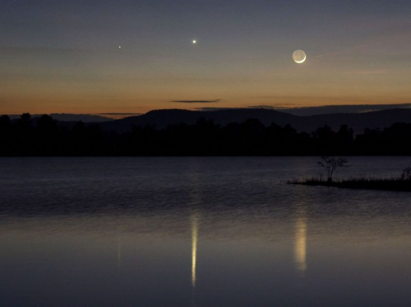 A trio of objects at twilight, whose reflection shines in a lake: fainter Mercury, brighter Venus and a very thin waxing crescent moon.