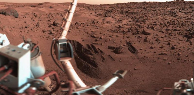 Sand drifts, rocks and reddish soil with narrow, deep trenches and robotic lander in foreground.