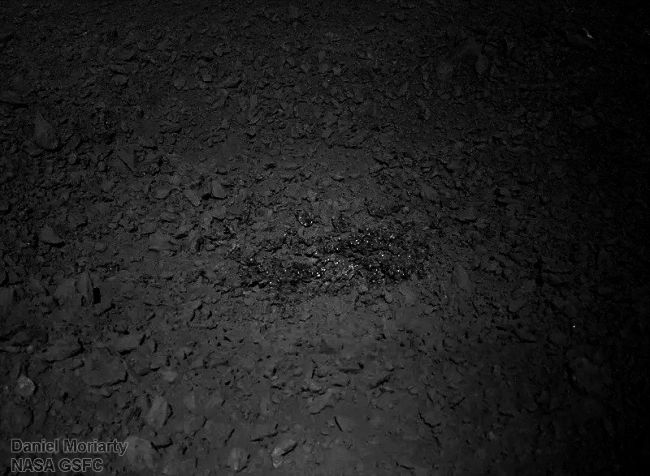 Dark view of gray pebbly soil with bright specks clustered in the middle.