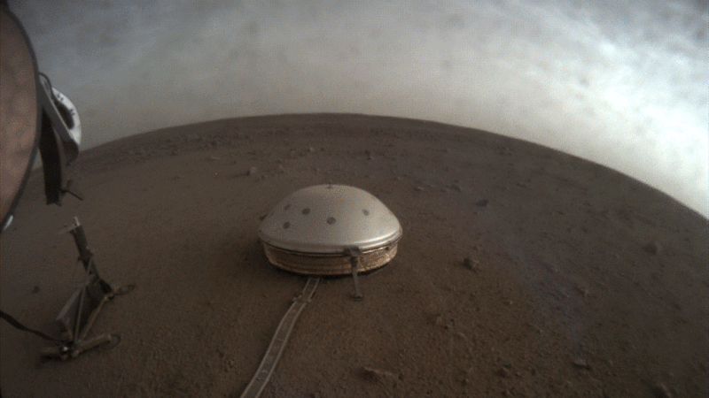 Gray dome at the end of a long flat cable on gray sandy surface with clouds flying overhead.