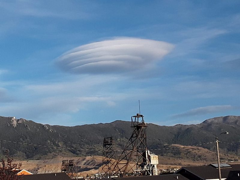 Flying saucer like cloud in blue sky over gray mountains with rusty mining equipemnt in foreground.