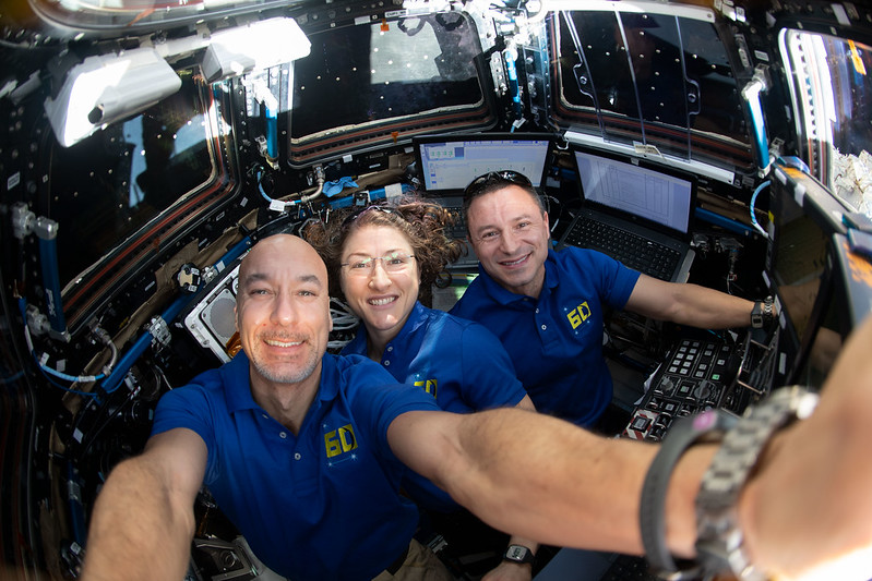 Three astronauts in blue shirts in small round chamber with three windows visible.