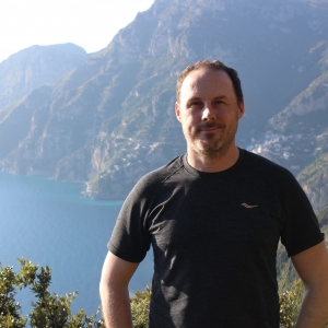 Man wearing t-shirt standing in front of mountain and shoreline.