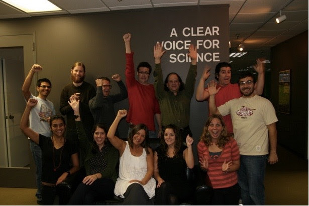 A group of jubilant people, sitting and standing under a sign that says 'A Clear Voice for Science'.