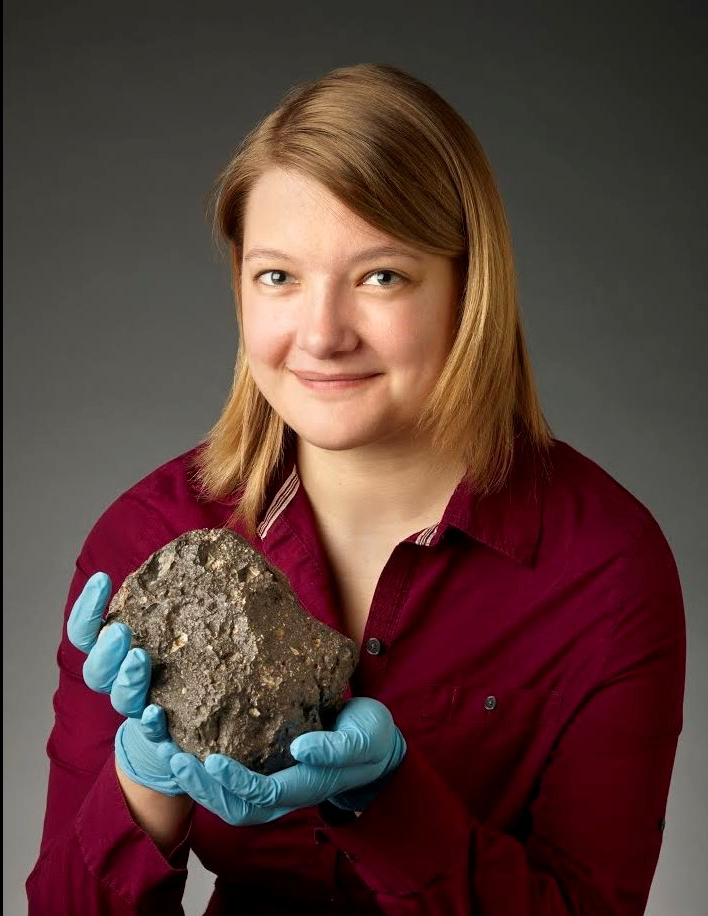 A smiling young woman wearing plastic gloves, holding a big rock.