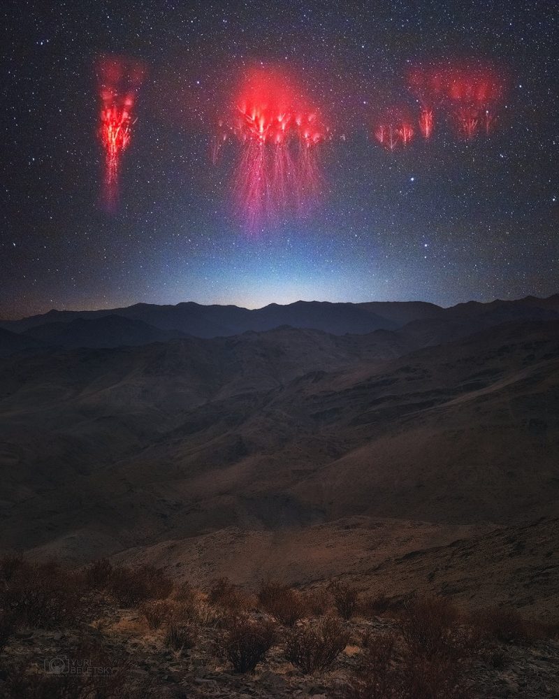 Dark sky with 3 fluorescent pink blobs wiht red lightning trailing down from them over mountains.