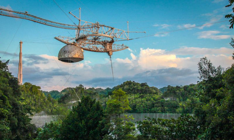 A view from the ground of the Arecibo Observatory dish, surrounded by thick forest, and receiver suspended above it.