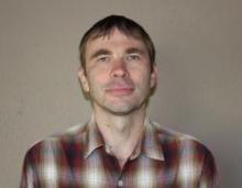 Dark-haired man with checkered shirt on light background.