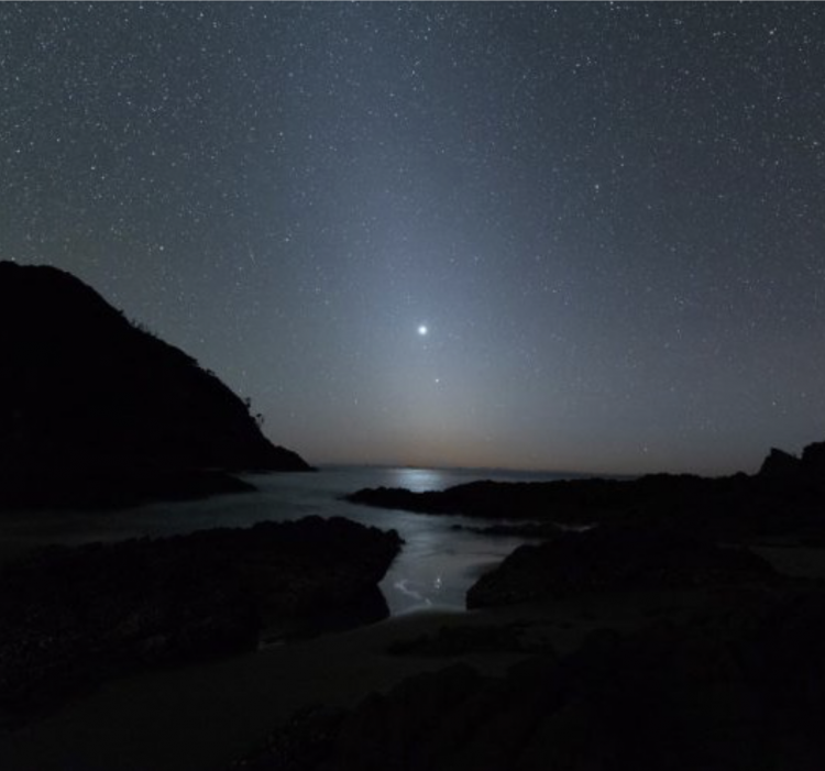 Fuzzy cone of light up from horizon in dark sky, with 2 bright dots, above rocky shoreline.