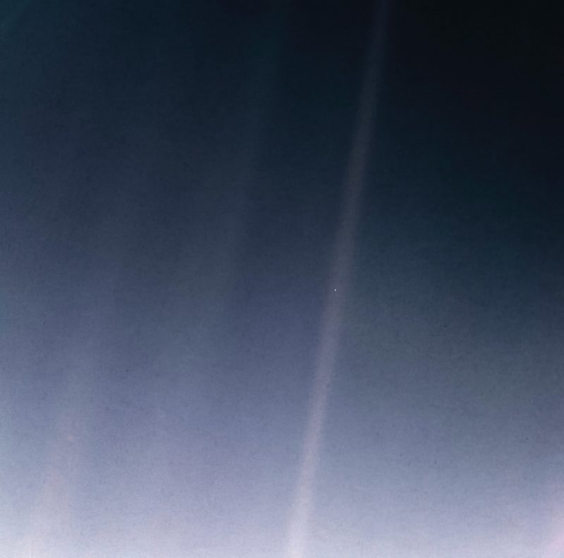 An image of bluish space, with streaks of sunlight crossing it, and with a single dot - Earth - within one of the sunbeams.