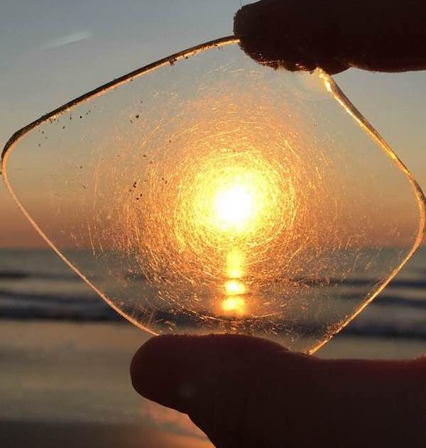 Two fingers holding up a rounded piece of glass with a sun setting over water behind.