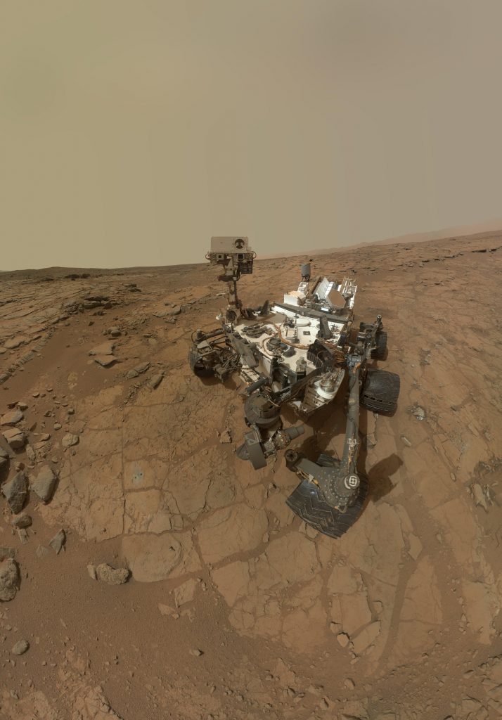 Mechanical rover with wheels and tools on extended arms, sitting on brownish rocky ground with dusty sky.