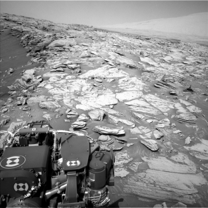 Gray rocky terrain with part of mechanical rover in foreground.